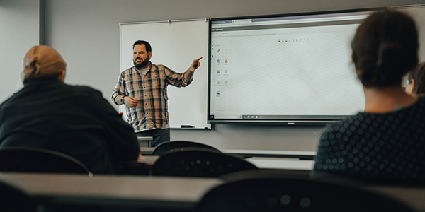 Professor standing in front of digital screen, presenting to a class.